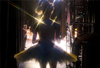 Accompanying photo of a dancer in a tutu seen from behind as she stands in the wings looking onto the stage